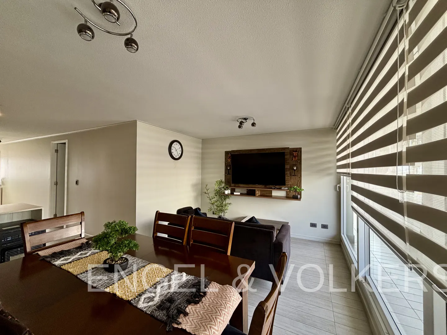 Living room with a dark wood dining table, chairs, and a brown sofa. A TV is mounted on the wall. Large windows with striped blinds.
