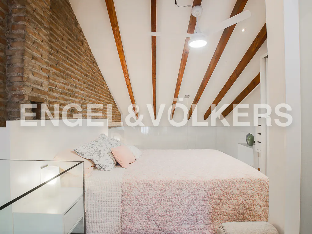 Bedroom with exposed brick wall, white bed, and wooden beams on the ceiling. A ceiling fan is visible.