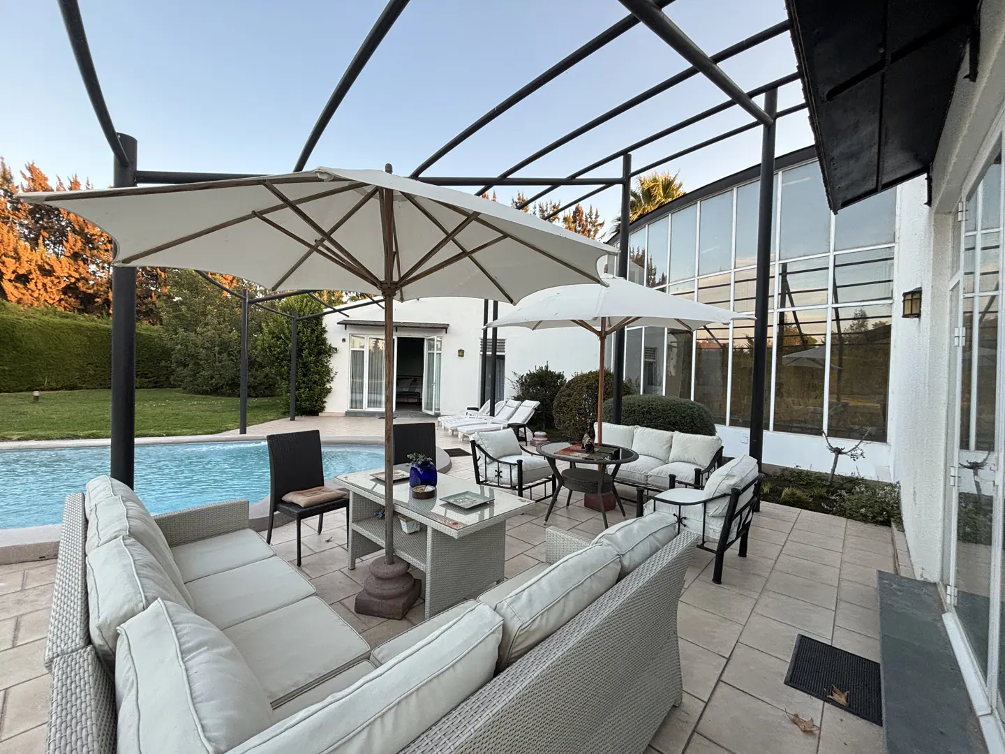 Outdoor patio with white wicker furniture, umbrellas, and a pool. A modern white house with large windows is in the background.