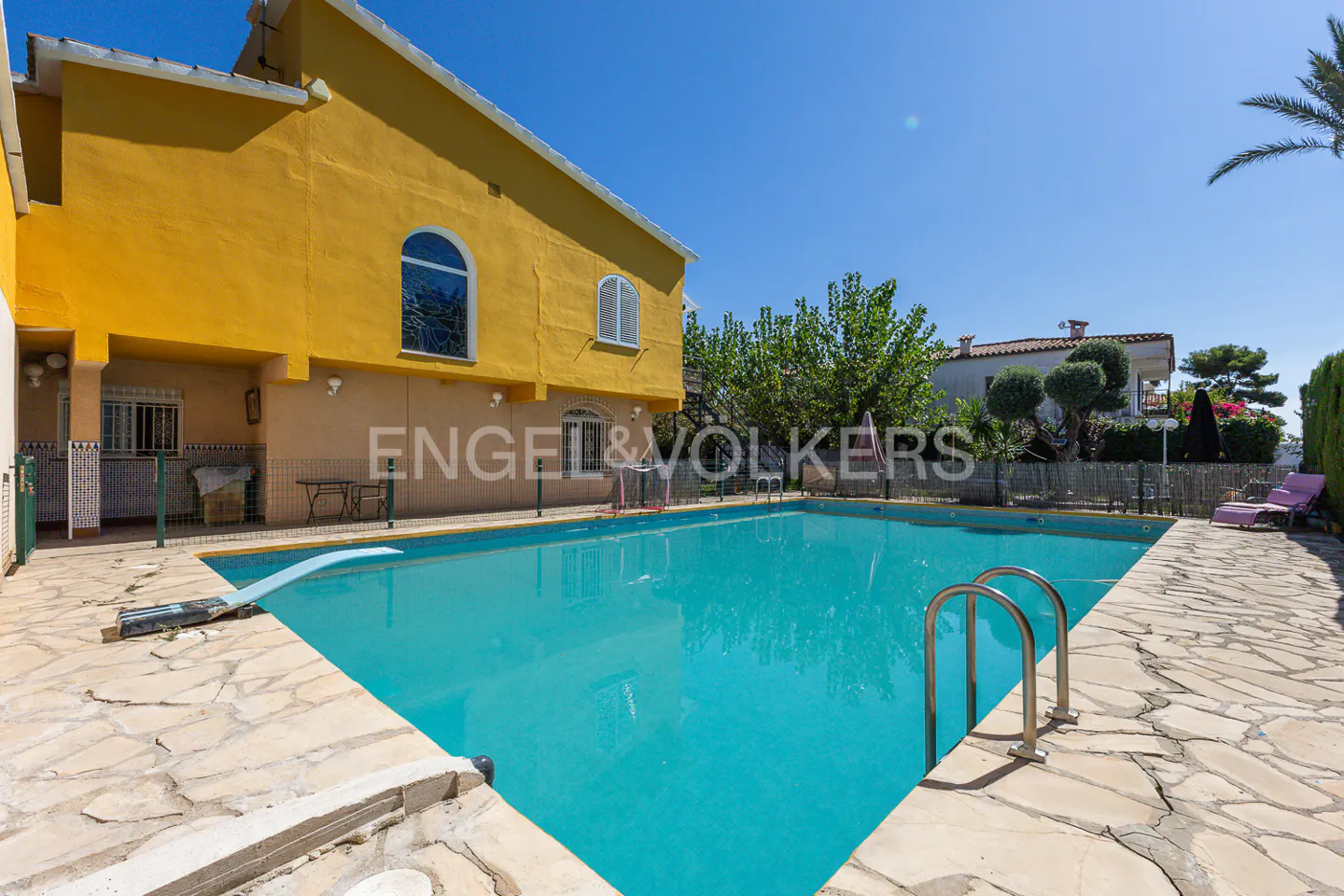 A yellow two-story house with a blue pool and stone patio under a clear blue sky.