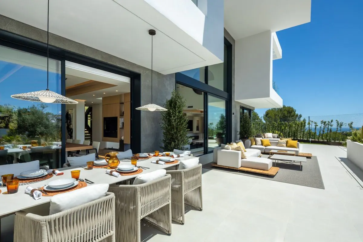 Modern outdoor patio with a dining table, woven chairs, and a lounge area under a white architectural overhang.
