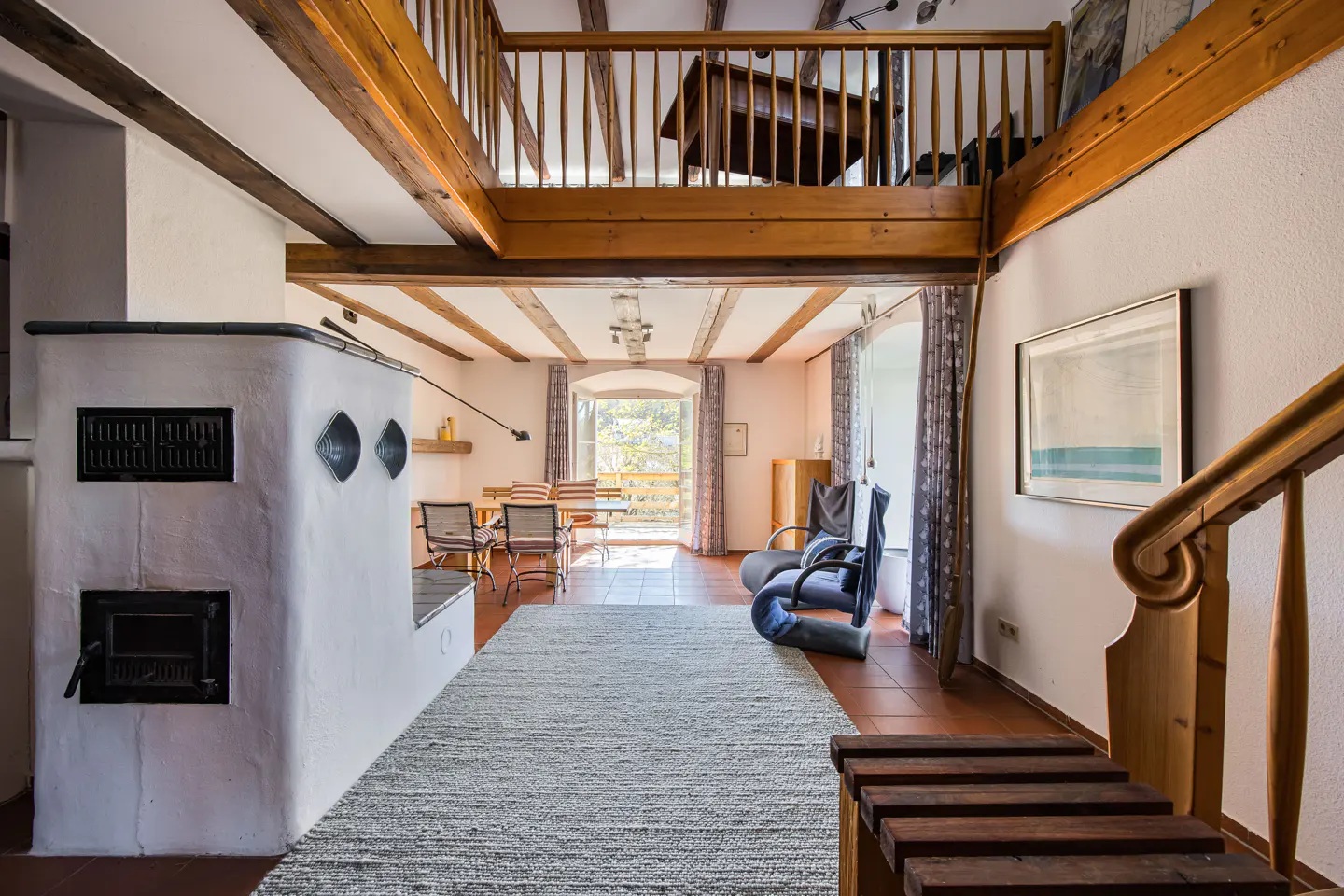 Interior view of a home with exposed wood beams, a white fireplace, and a dining area leading to a balcony. A staircase is visible on the right.