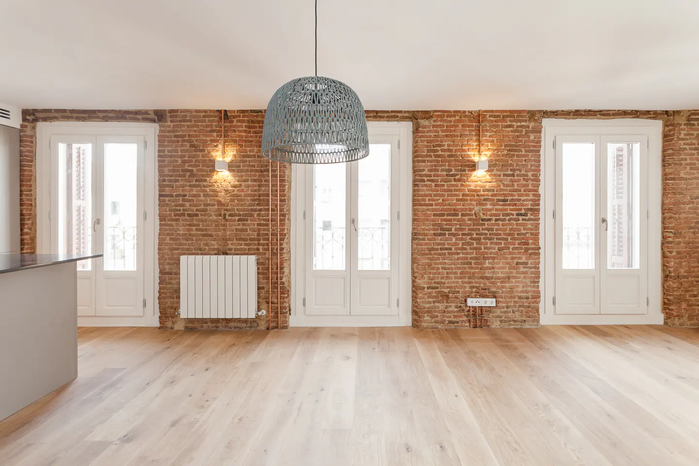 Empty room with hardwood floors, brick wall, and three white doors. A gray woven pendant light hangs from the ceiling.