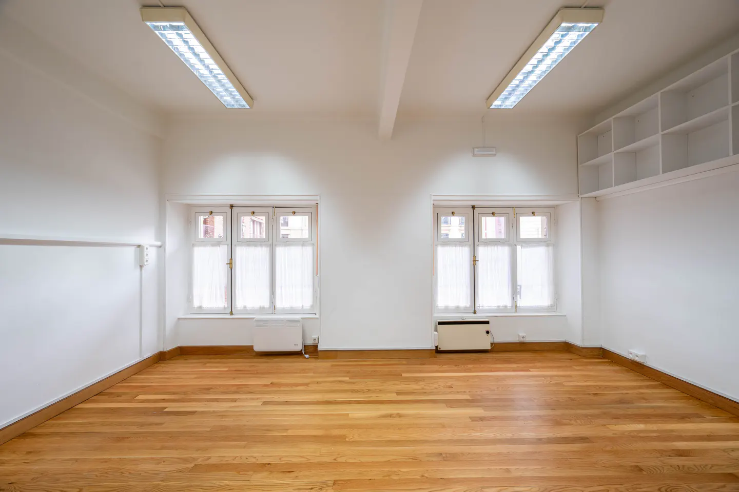 Bright, empty room with hardwood floors, white walls, two windows with sheer curtains, and fluorescent lighting.