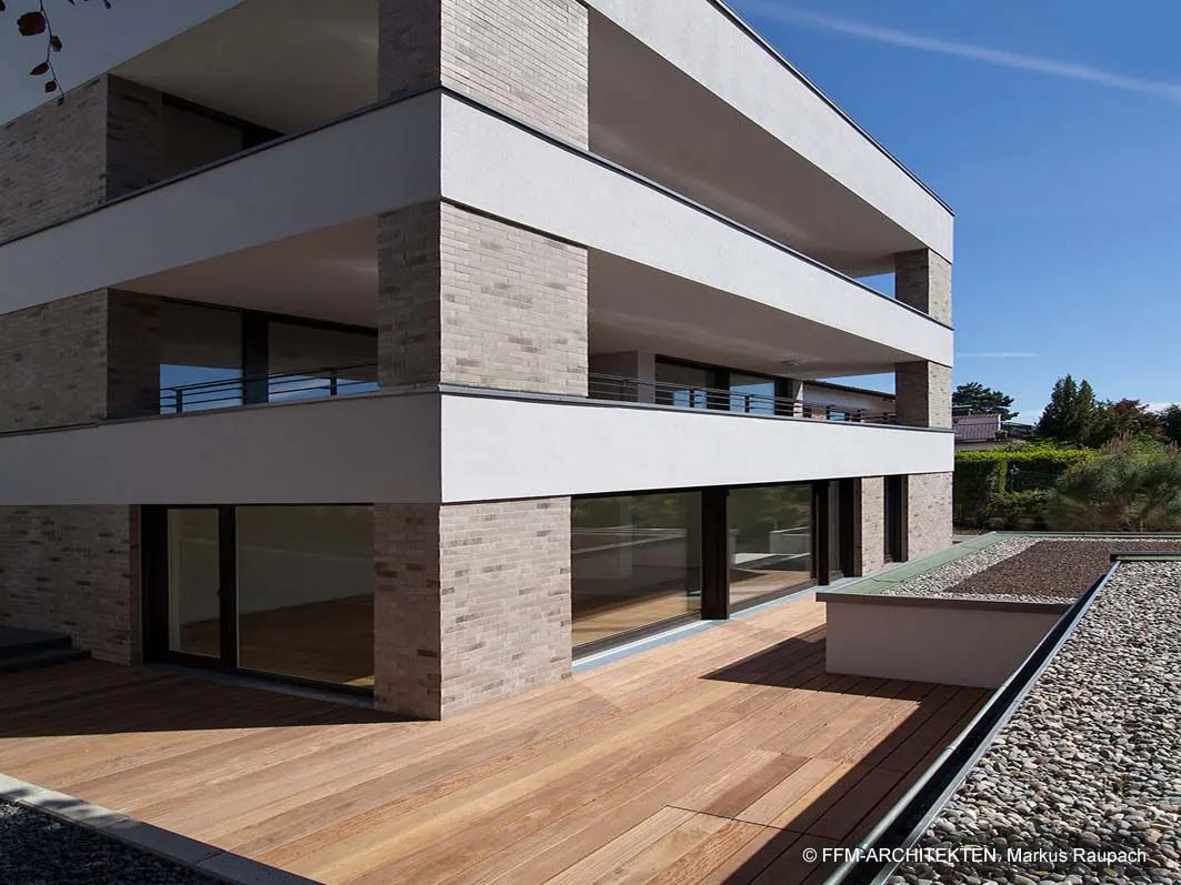 Modern building exterior with a wood deck, gray brick accents, white trim, and large windows under a blue sky.