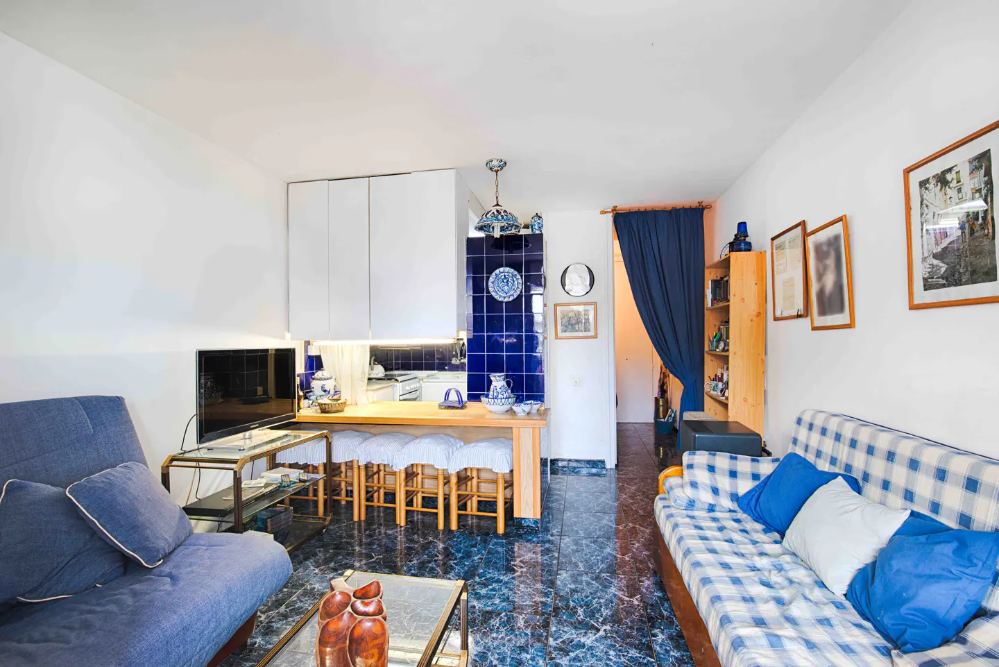 A cozy living room with blue marble floors, a blue sofa, and a blue-and-white plaid sofa. A kitchen area with white cabinets and blue tile backsplash is visible.