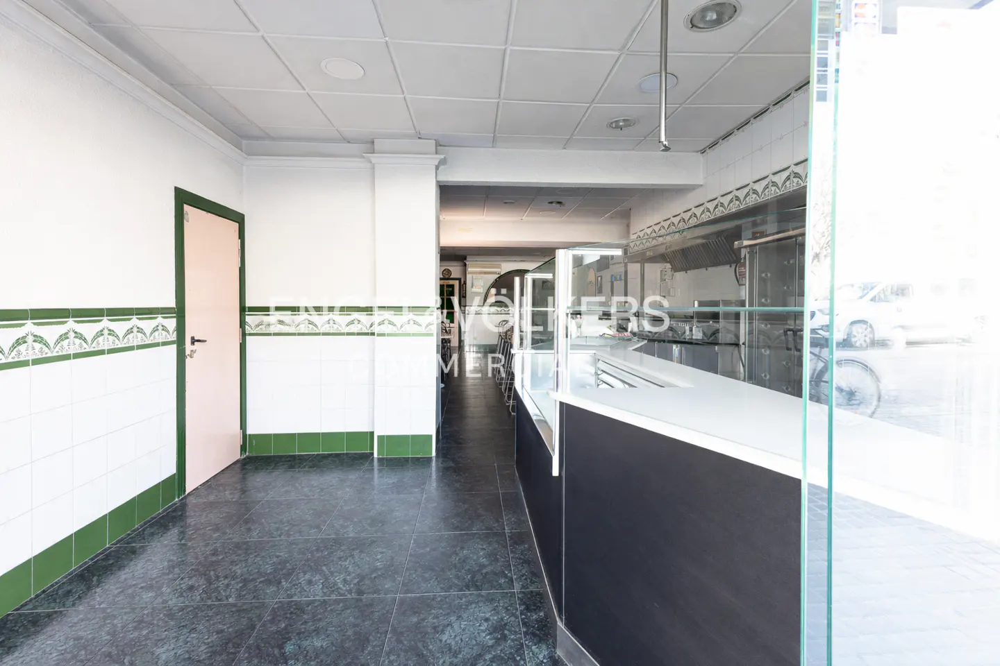 Empty commercial space with white walls, green trim, and dark marble floors. A counter runs along the right side, and a door is on the left.