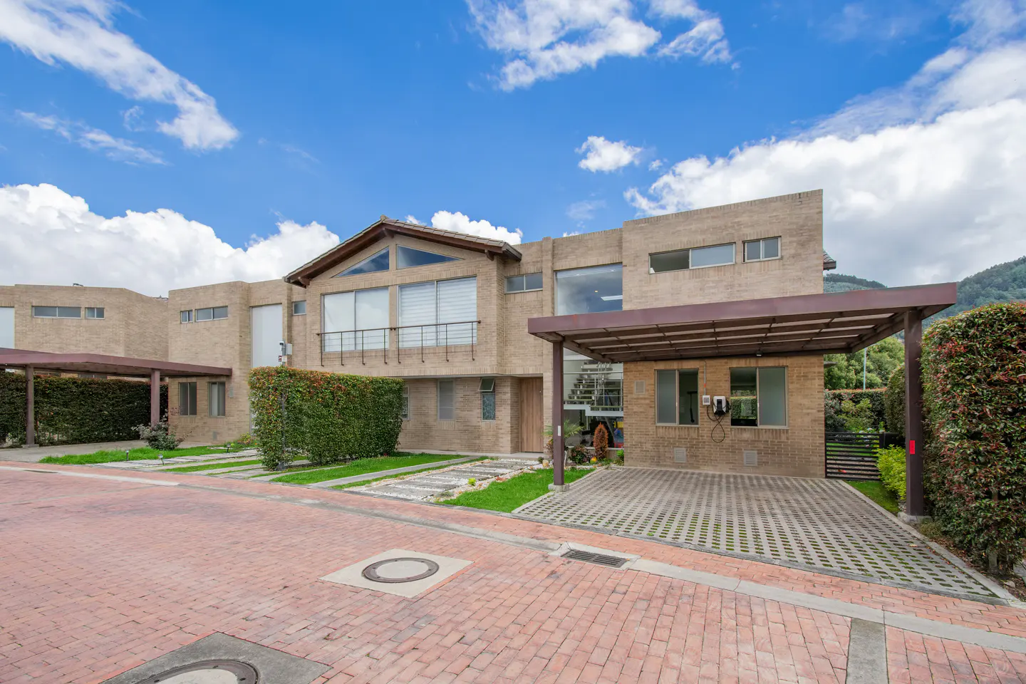 Exterior view of a modern, tan brick house with a carport, green hedges, and a red brick driveway under a blue sky.