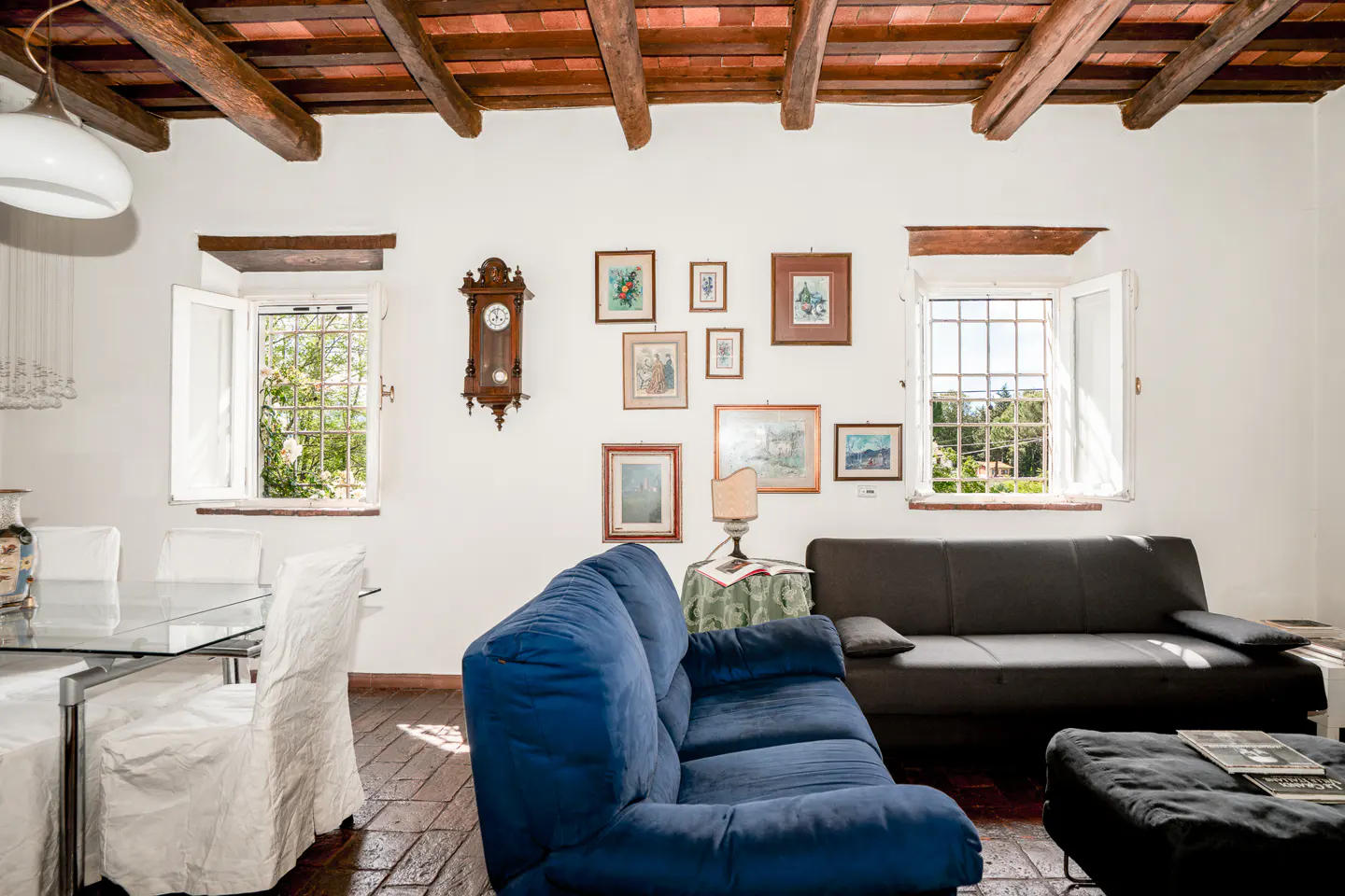 Living room with exposed wood ceiling, blue sofa, black couch, dining area, and a gallery wall of framed art on white walls.