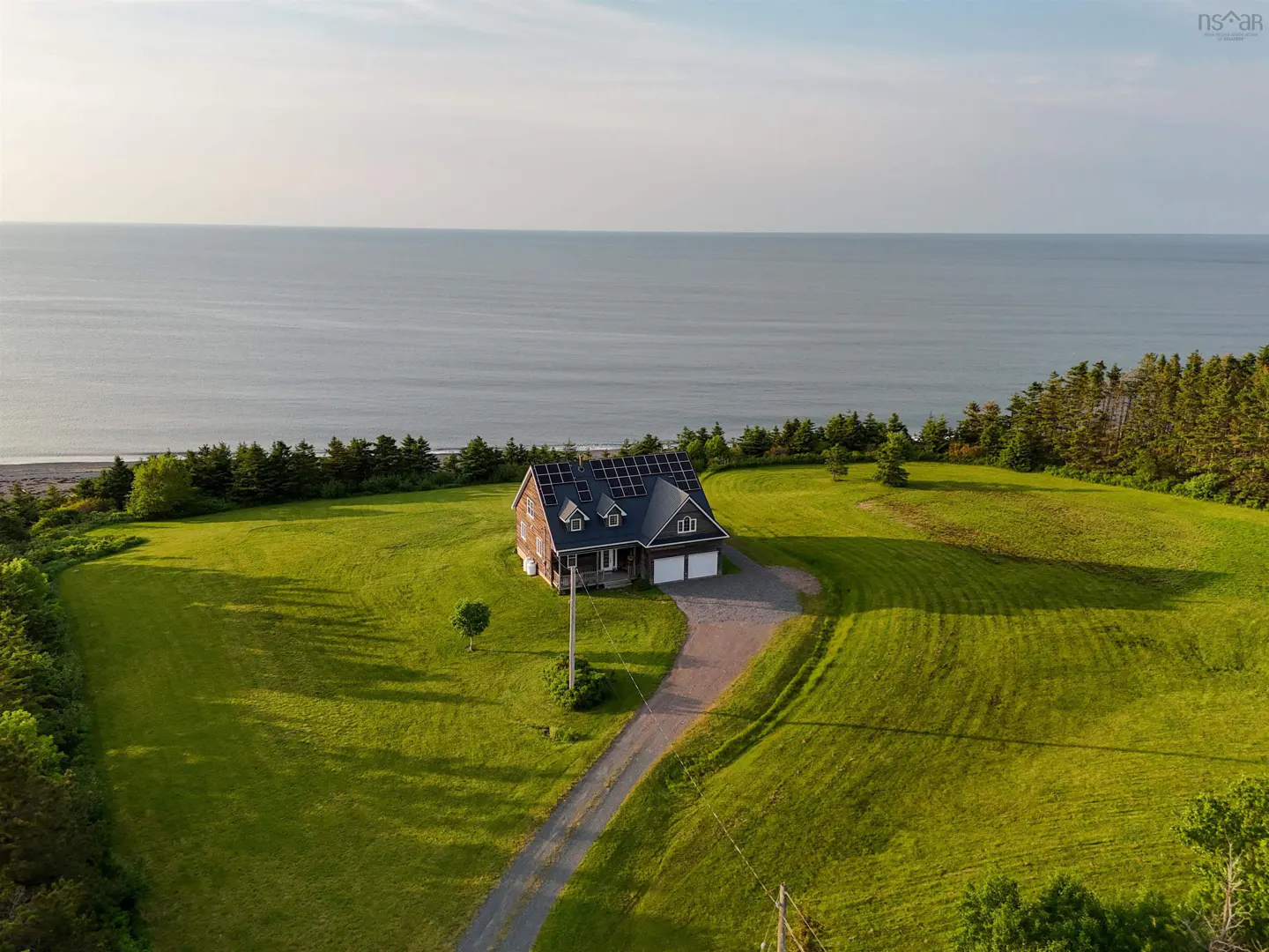 Aerial view of a brown house with solar panels, a green lawn, and the ocean in the background.
