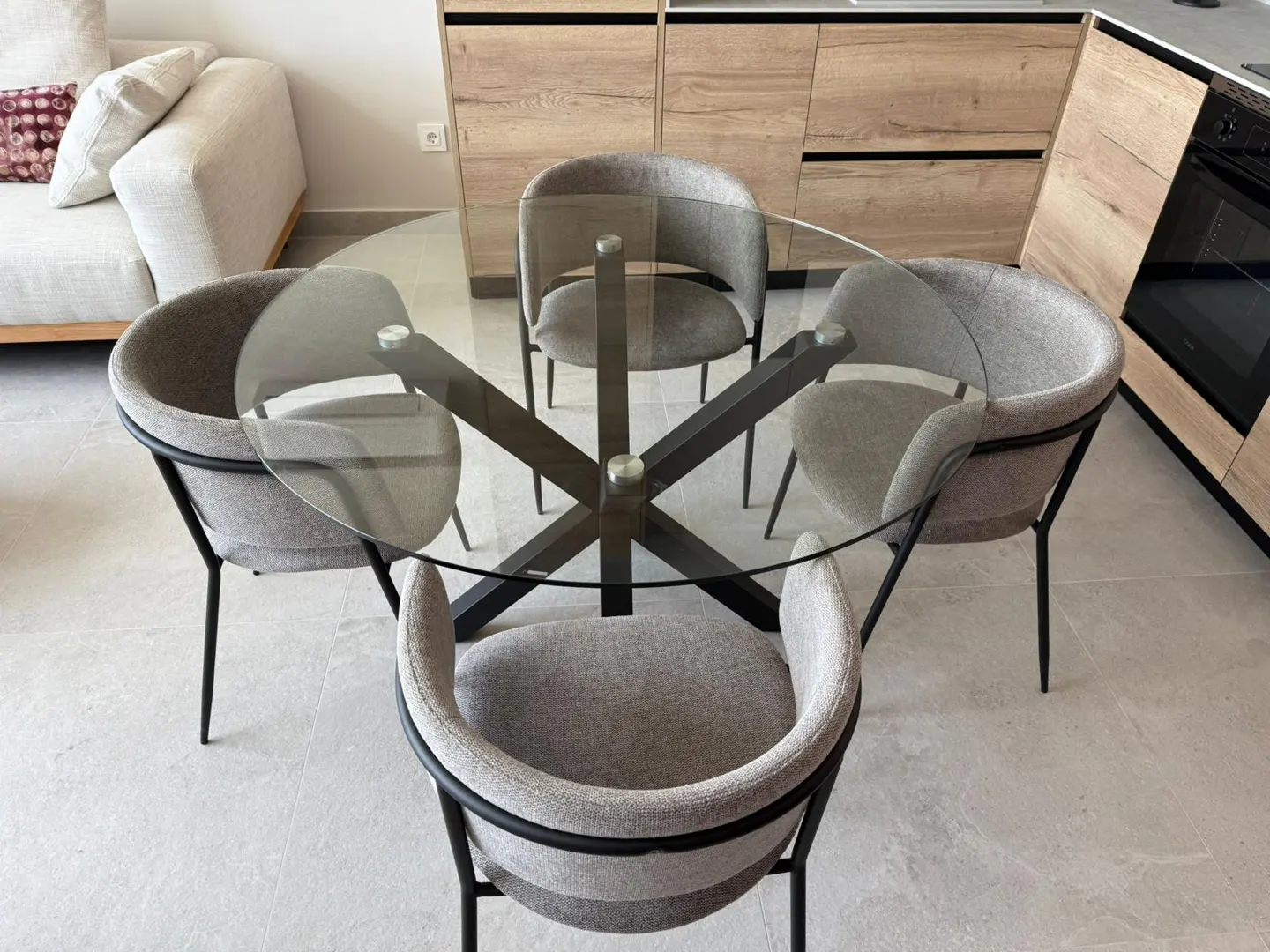 A modern kitchen features a round glass table with gray chairs and a black base, complemented by light wood cabinets.