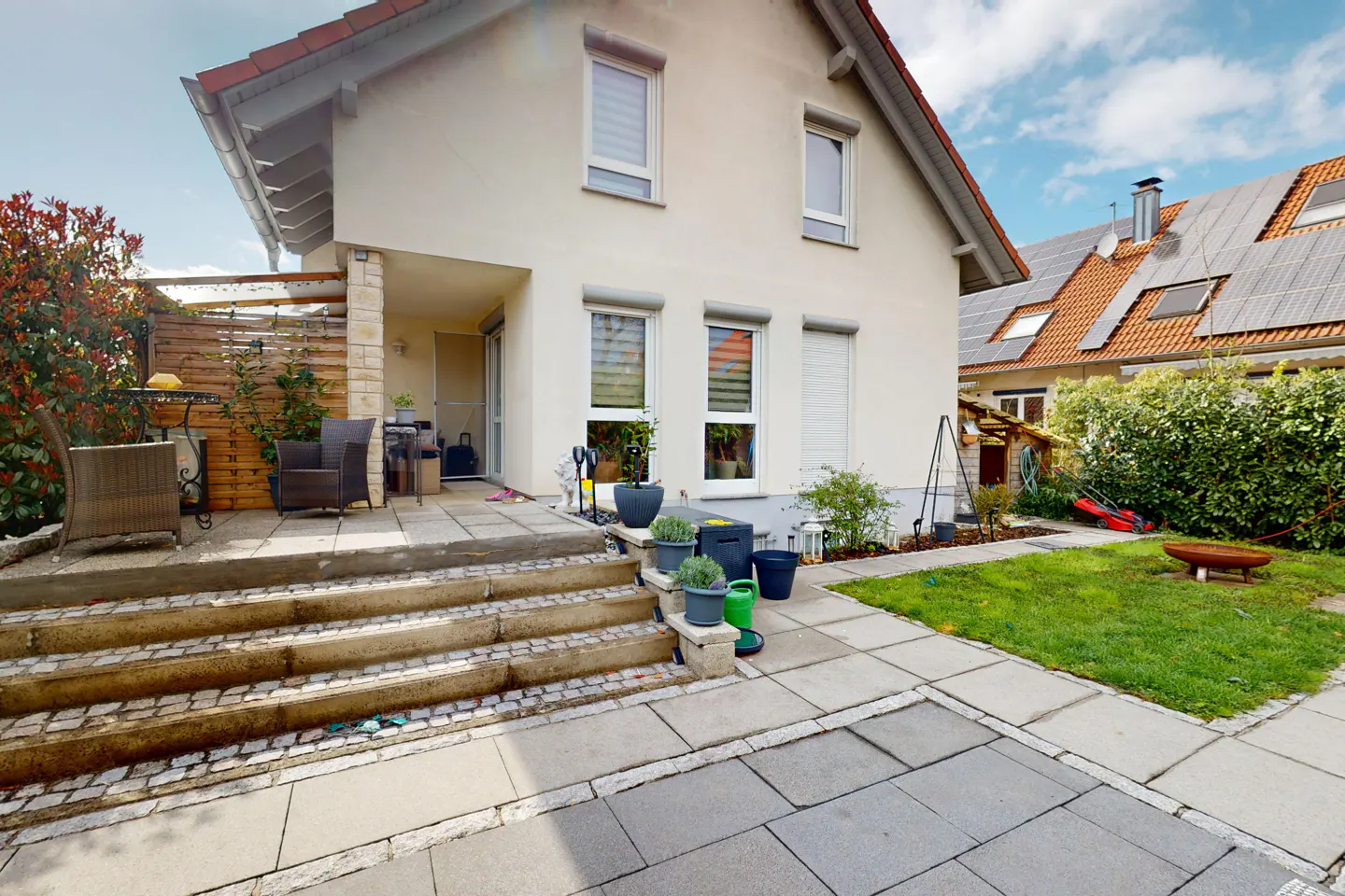 Exterior view of a two-story house with a patio, stone steps, and a small lawn. The house is painted in a light color with a red tile roof.