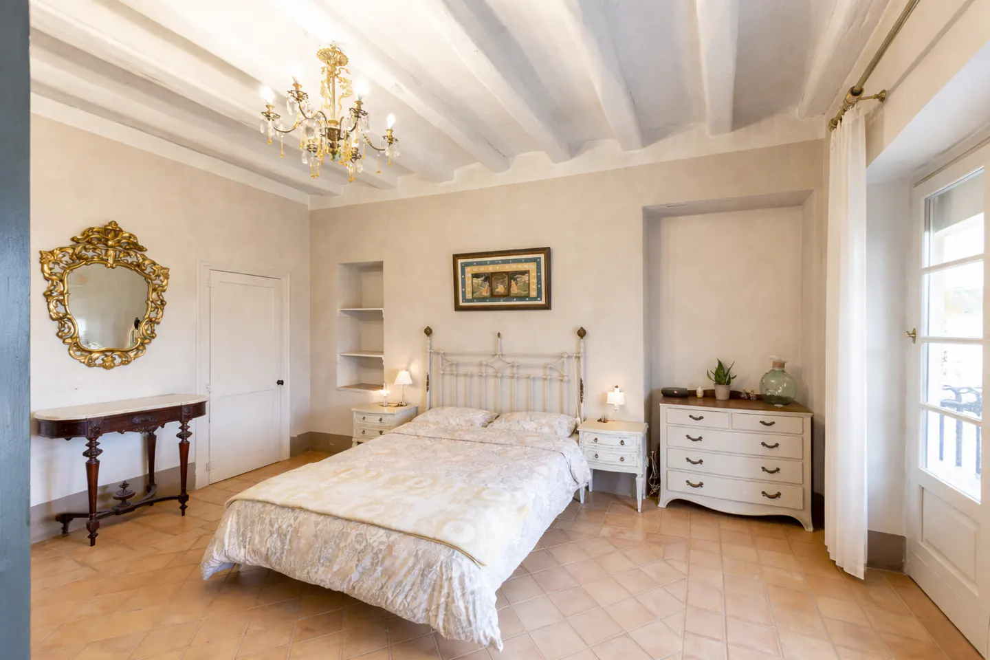 Bedroom with white walls, terracotta tile floor, and white beamed ceiling. A wrought iron bed is centered, flanked by white nightstands. A gold mirror hangs above a dark wood table.