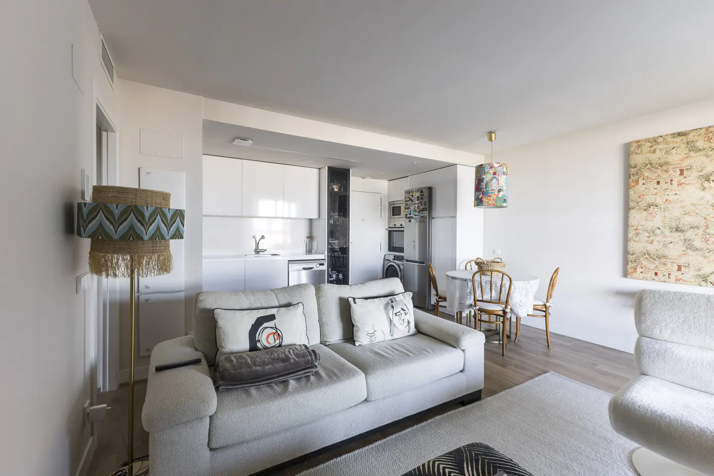 Bright, modern apartment interior with a white sofa, kitchen, and dining area. A unique floor lamp and artwork add character.