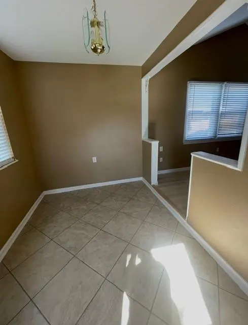 A tan-walled room with tile floors, a gold chandelier, and a white-framed opening to another room with a window.