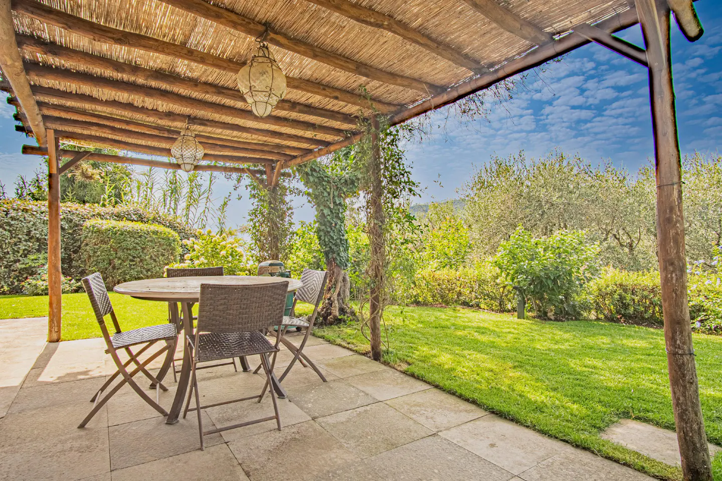Outdoor patio with a round table and four chairs under a thatched roof pergola, overlooking a green lawn and garden.
