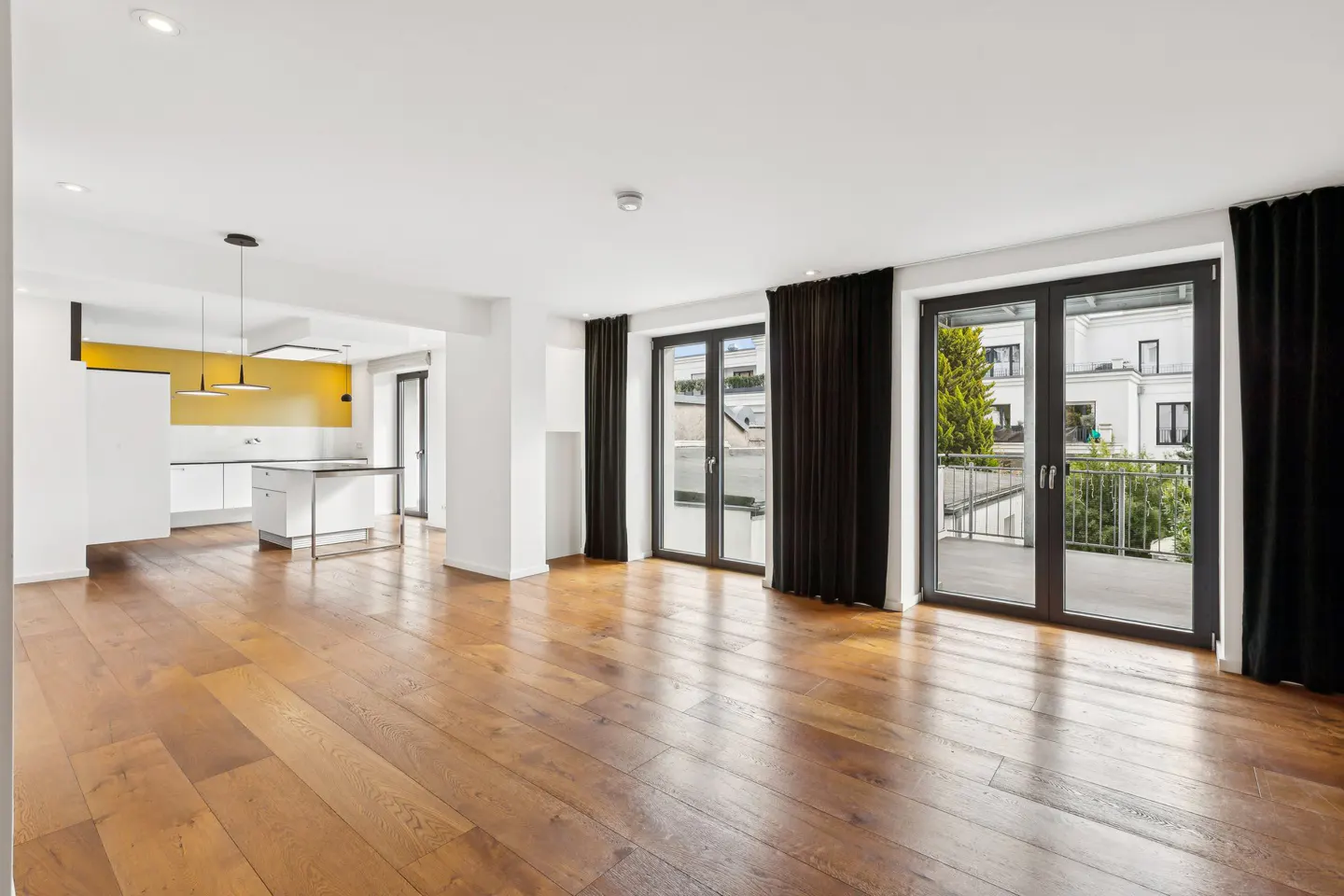 Bright, open-concept apartment with hardwood floors, white walls, and black-framed glass doors to a balcony. A modern kitchen with yellow accents is visible.