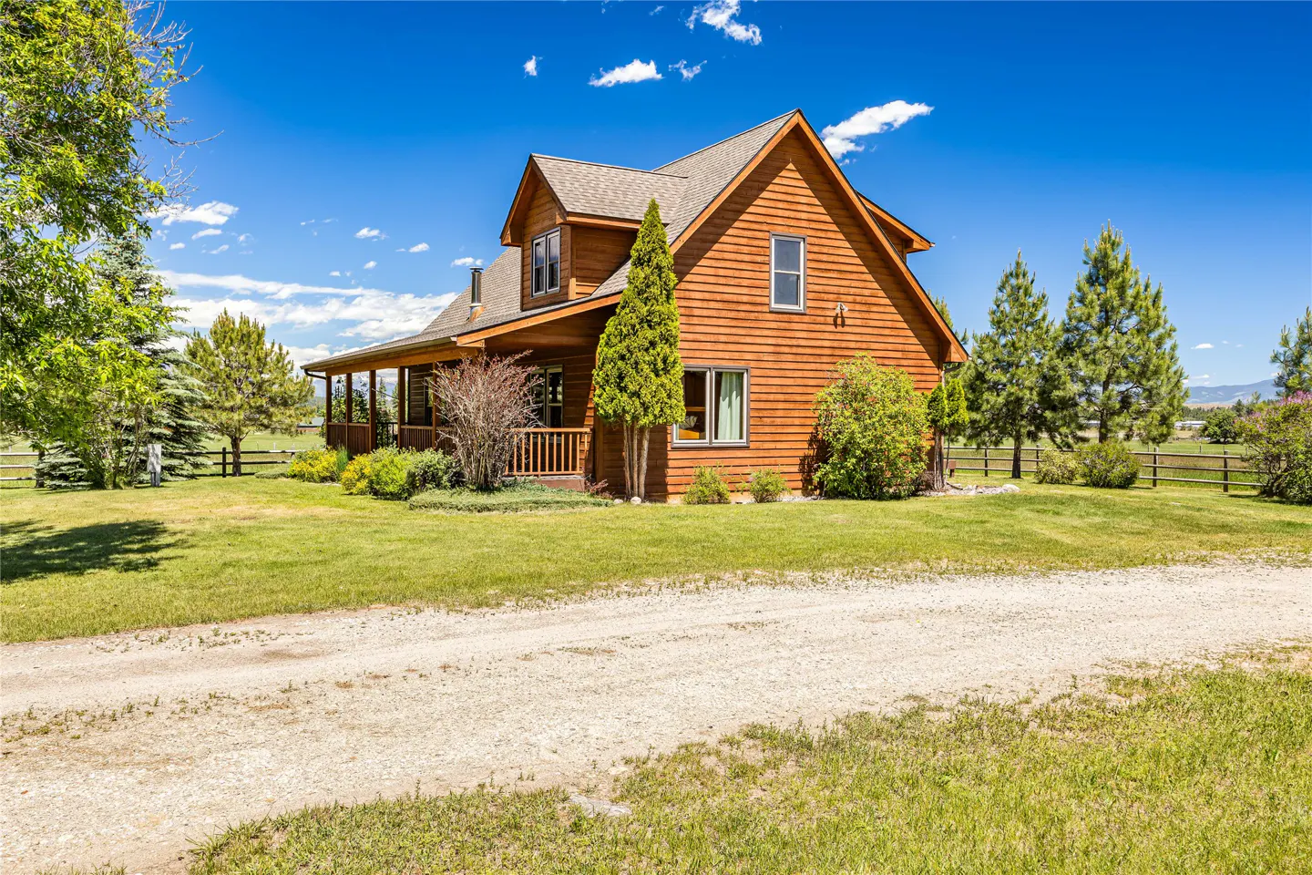 A two-story brown wood-sided cabin with a covered porch, surrounded by green lawns and pine trees under a blue sky.