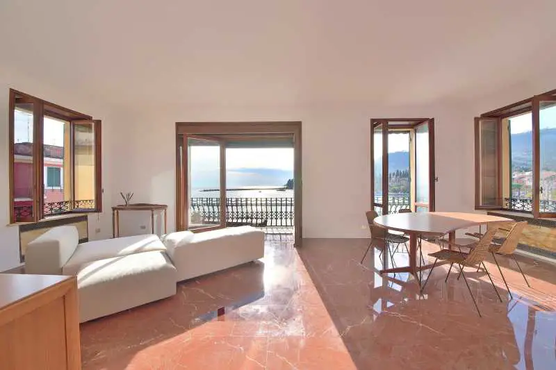 Bright living room with red marble floors, white sofa, and dining table. Balcony doors open to a sea view.