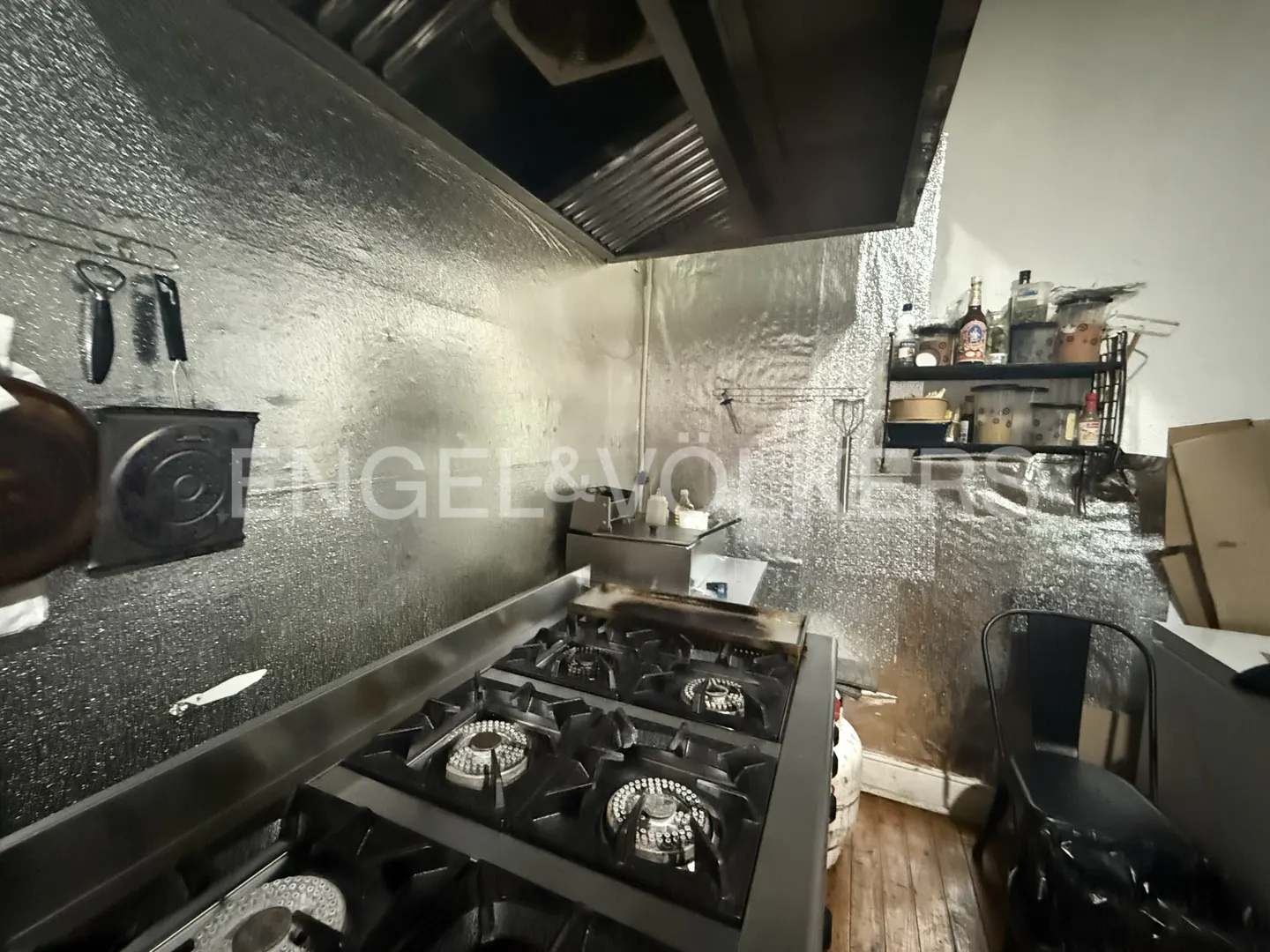 Commercial kitchen with a large stainless steel gas stove, metallic wall panels, a vent hood, and a black metal chair.