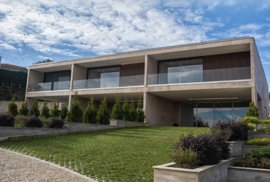 Modern two-story beige stone house with glass balconies, large windows, and a landscaped lawn under a blue cloudy sky.