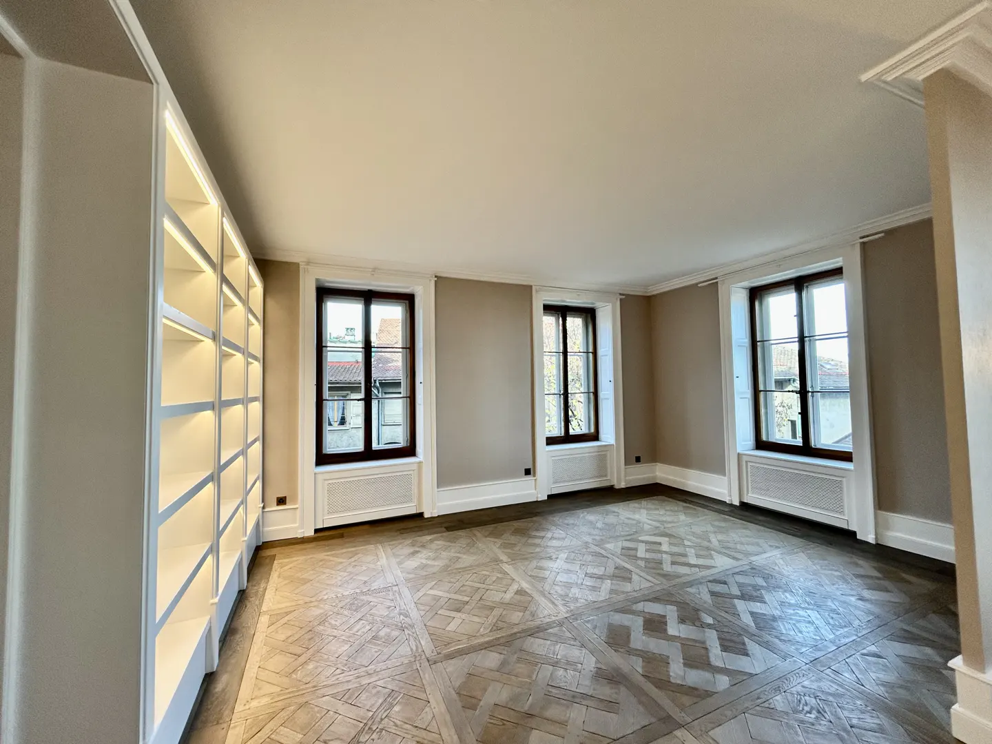 Bright, empty room with parquet floors, beige walls, and white trim. Three windows with dark frames and a built-in, illuminated bookshelf.