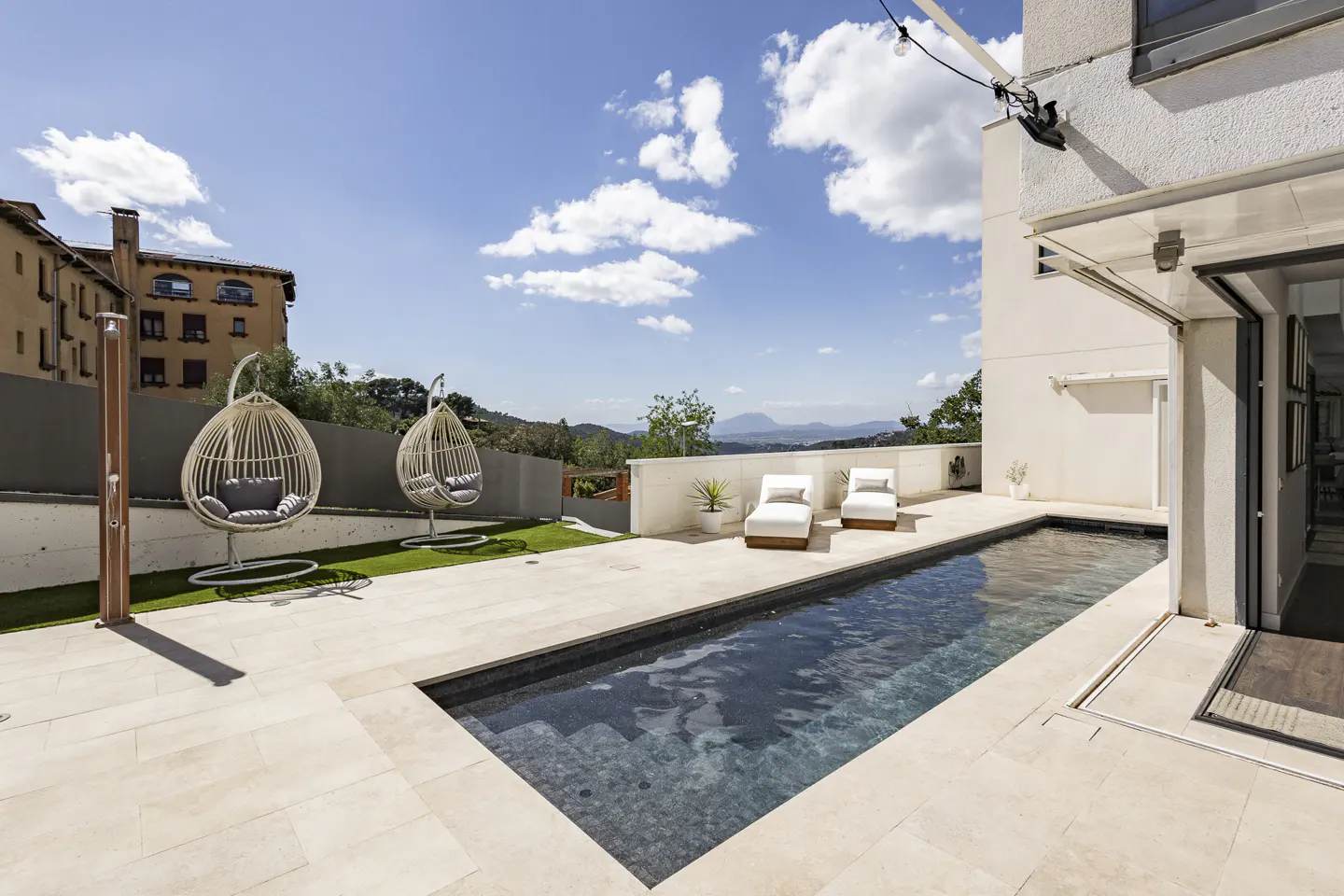 Outdoor patio with a narrow pool, two egg-shaped chairs, and two white lounge chairs. Blue sky with clouds in the background.