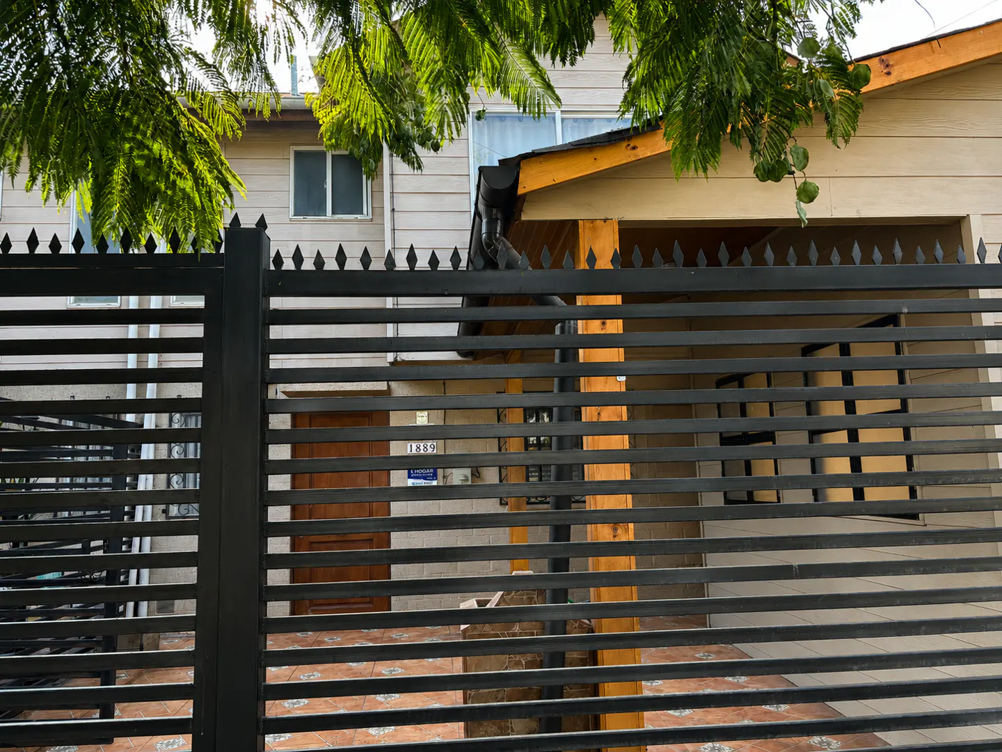 Two-story beige house with a wooden door and tiled patio, viewed through a black metal security fence.
