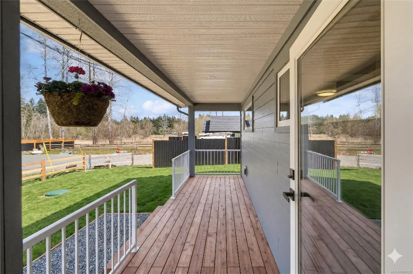 Covered wooden deck with white railings, a hanging flower basket, and a view of a grassy yard and rural landscape.