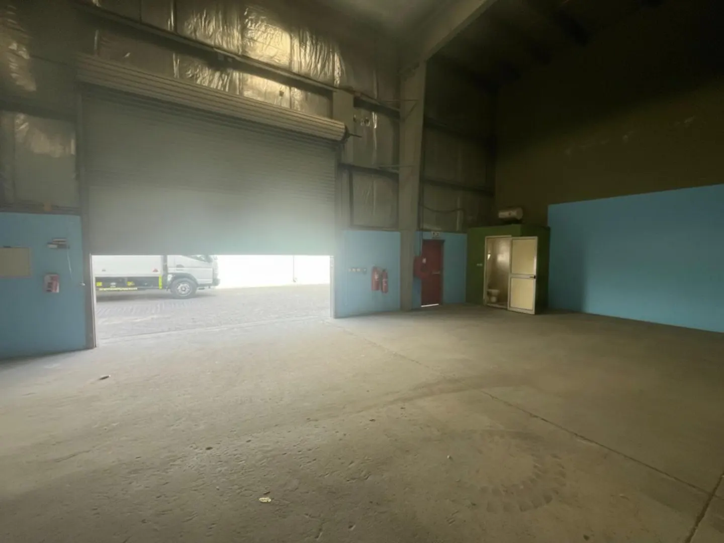 Interior view of a large, empty warehouse with a partially open roller door and a truck visible outside.