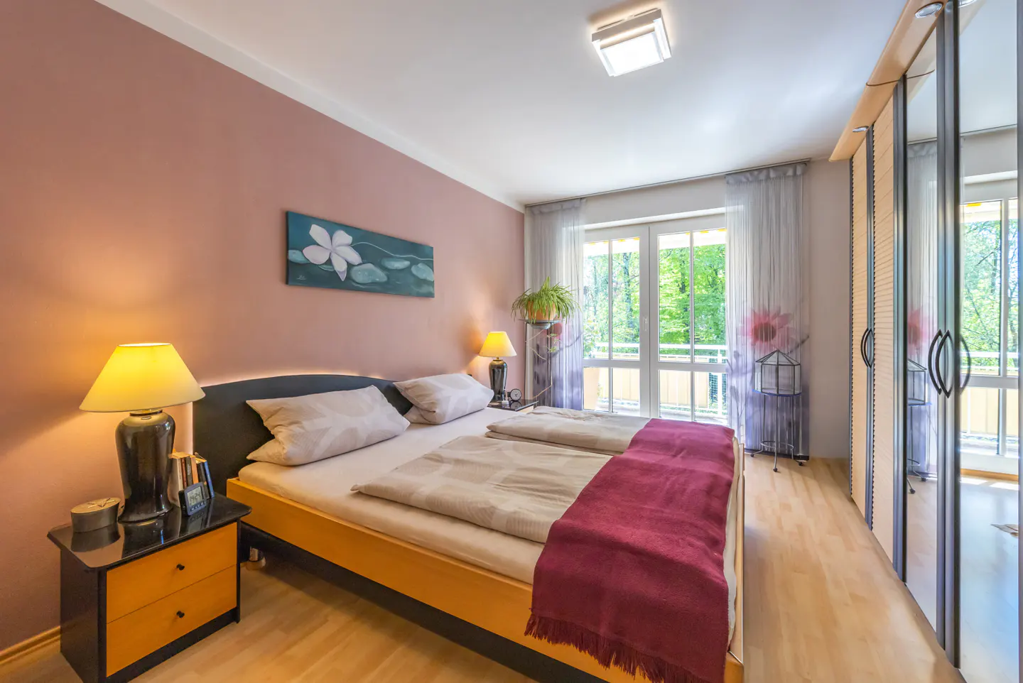 Bedroom with a double bed, pink wall, bedside table with lamp, and a balcony with a view of green trees.