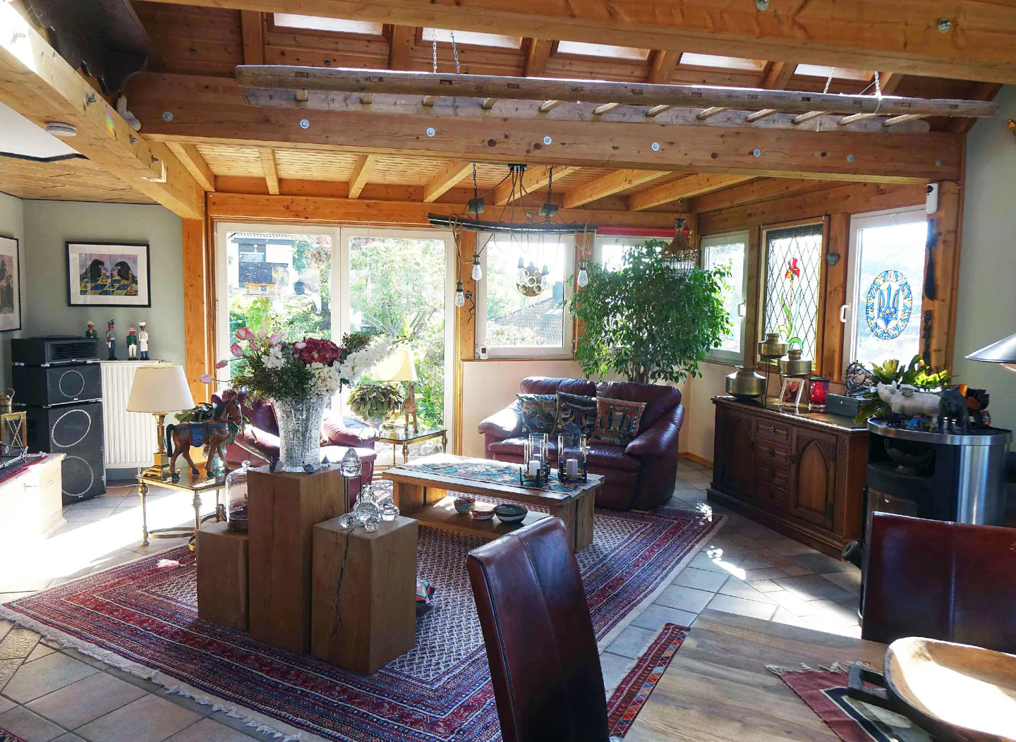 Sunlit living room with wood beams, ladder decor, leather furniture, and a patterned rug. Windows offer a view of greenery.