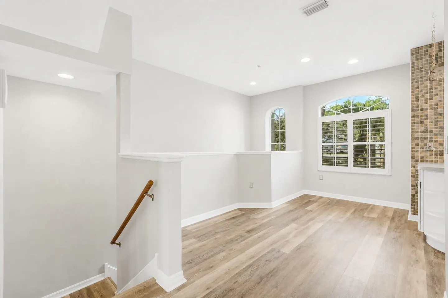 Bright, airy room with wood floors, white walls, and two arched windows with white shutters. A staircase with a wooden handrail is visible.