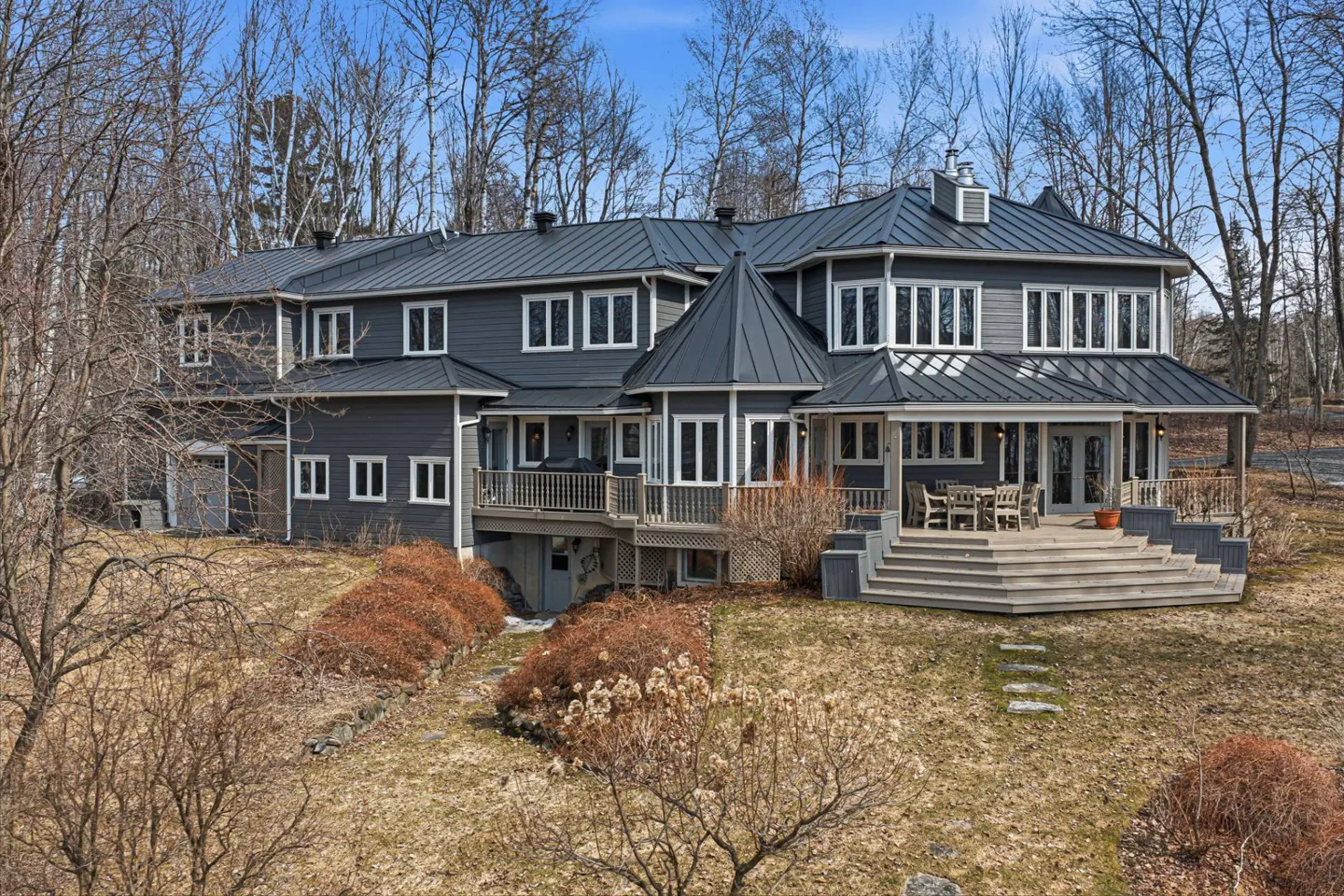 Two-story gray house with white trim and a metal roof, surrounded by trees and brown grass. A large porch with outdoor furniture is visible.