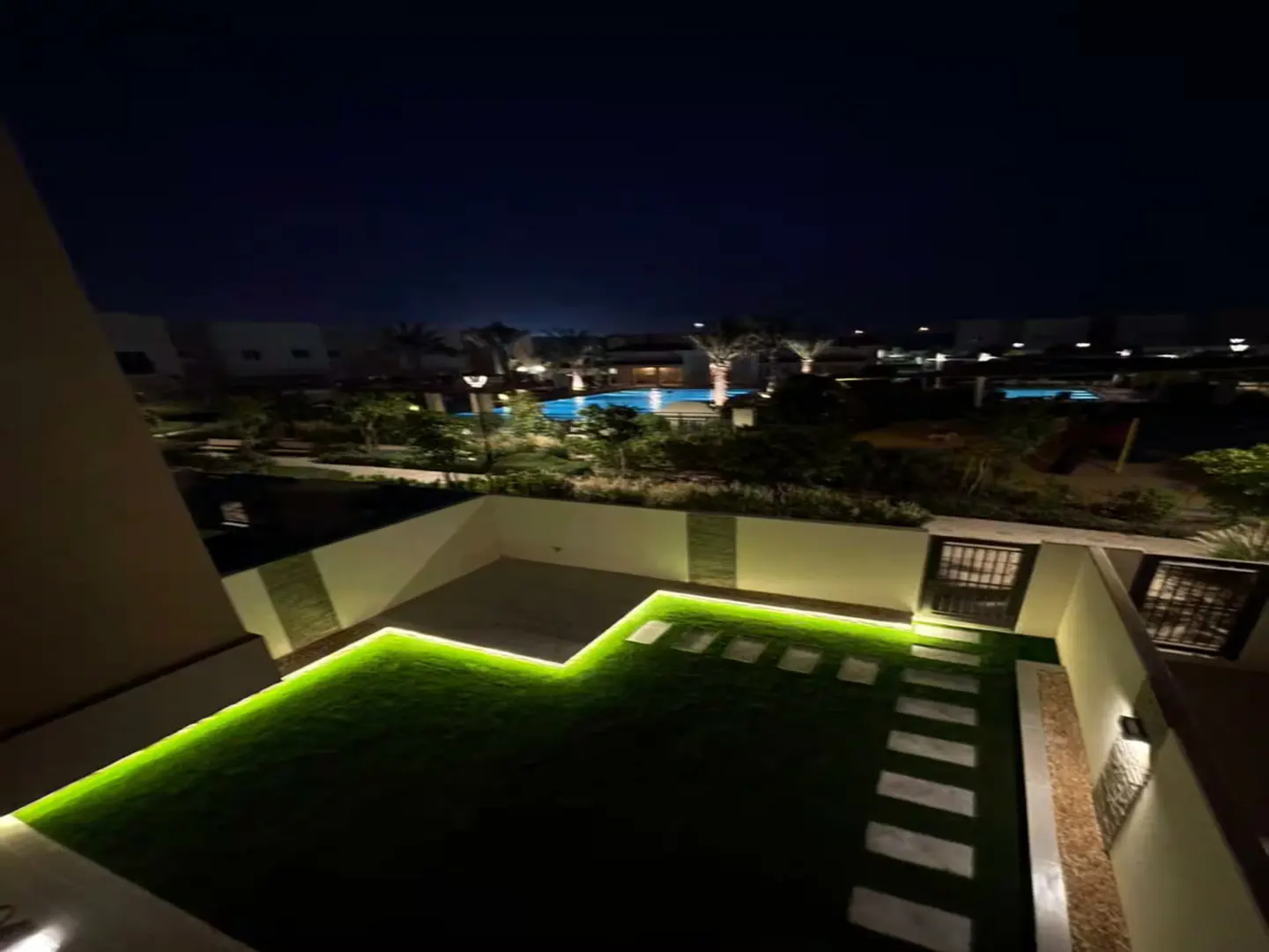 Night view of a backyard with green grass, stone path, and illuminated pool in the distance.