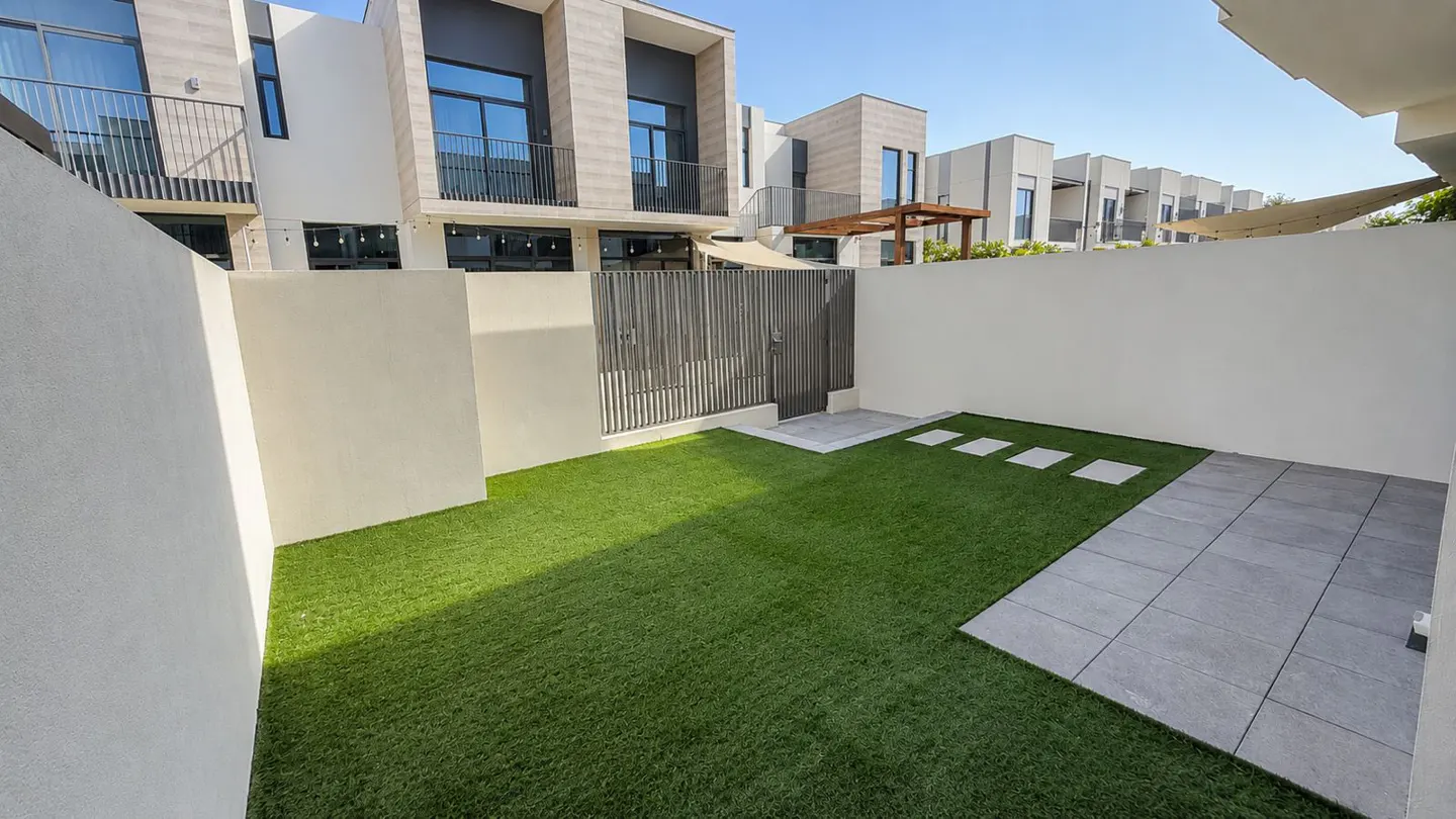 A backyard with green grass, a gray stone path, and a gray fence, surrounded by white walls and modern houses.