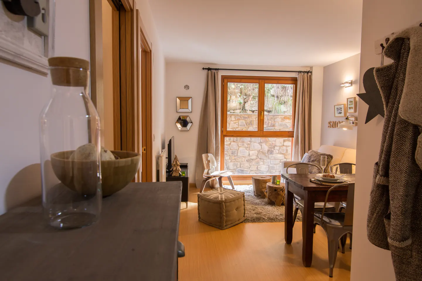 Cozy living room with wood floors, a dining table set for two, and a large window overlooking a stone wall.