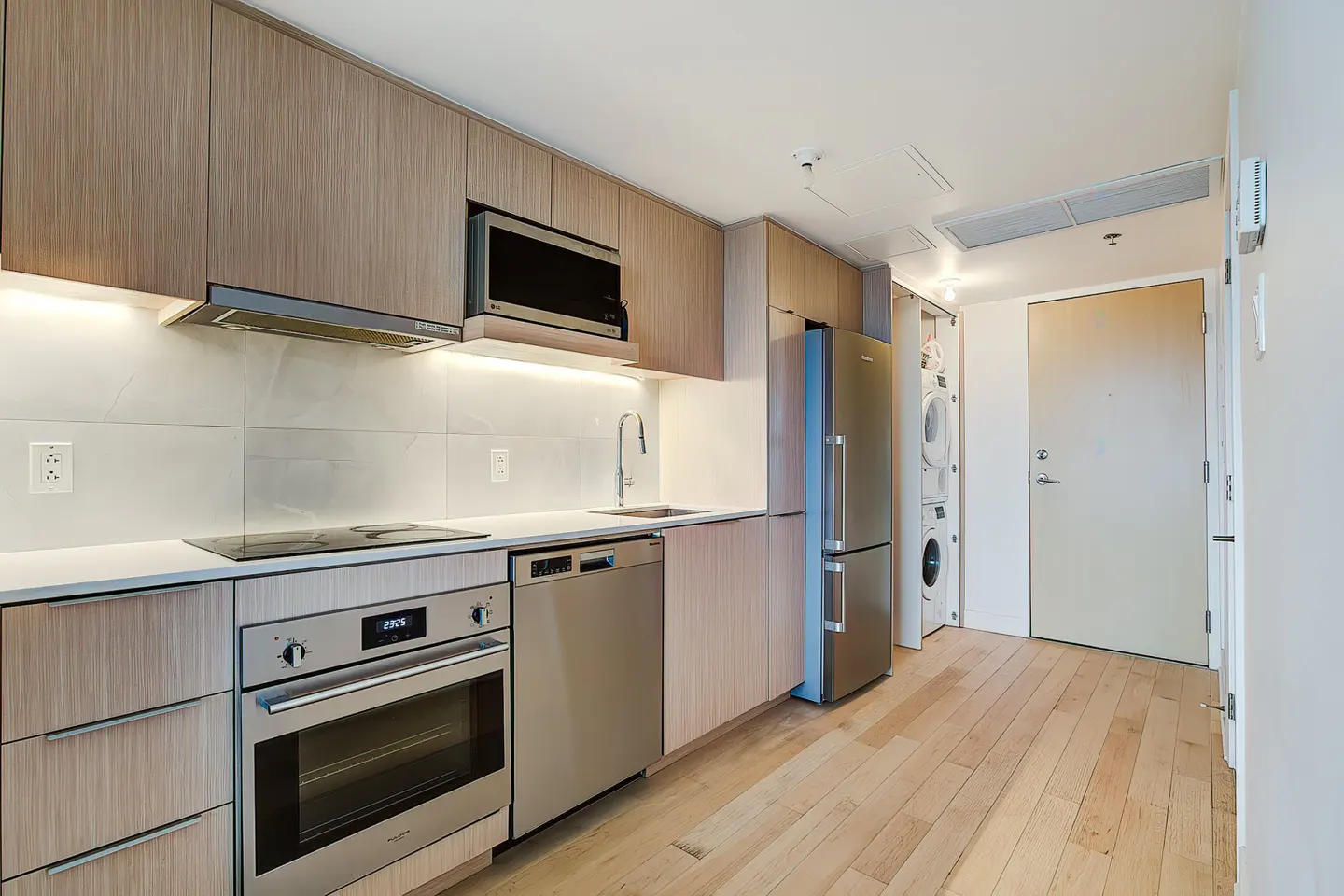 A modern kitchen with light wood cabinets, stainless steel appliances, and a white countertop. A washer and dryer are visible near the door.
