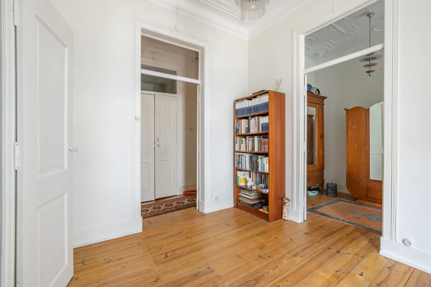 A bright hallway with wooden floors, white walls, and doorways. A tall bookcase stands against the wall, filled with books.