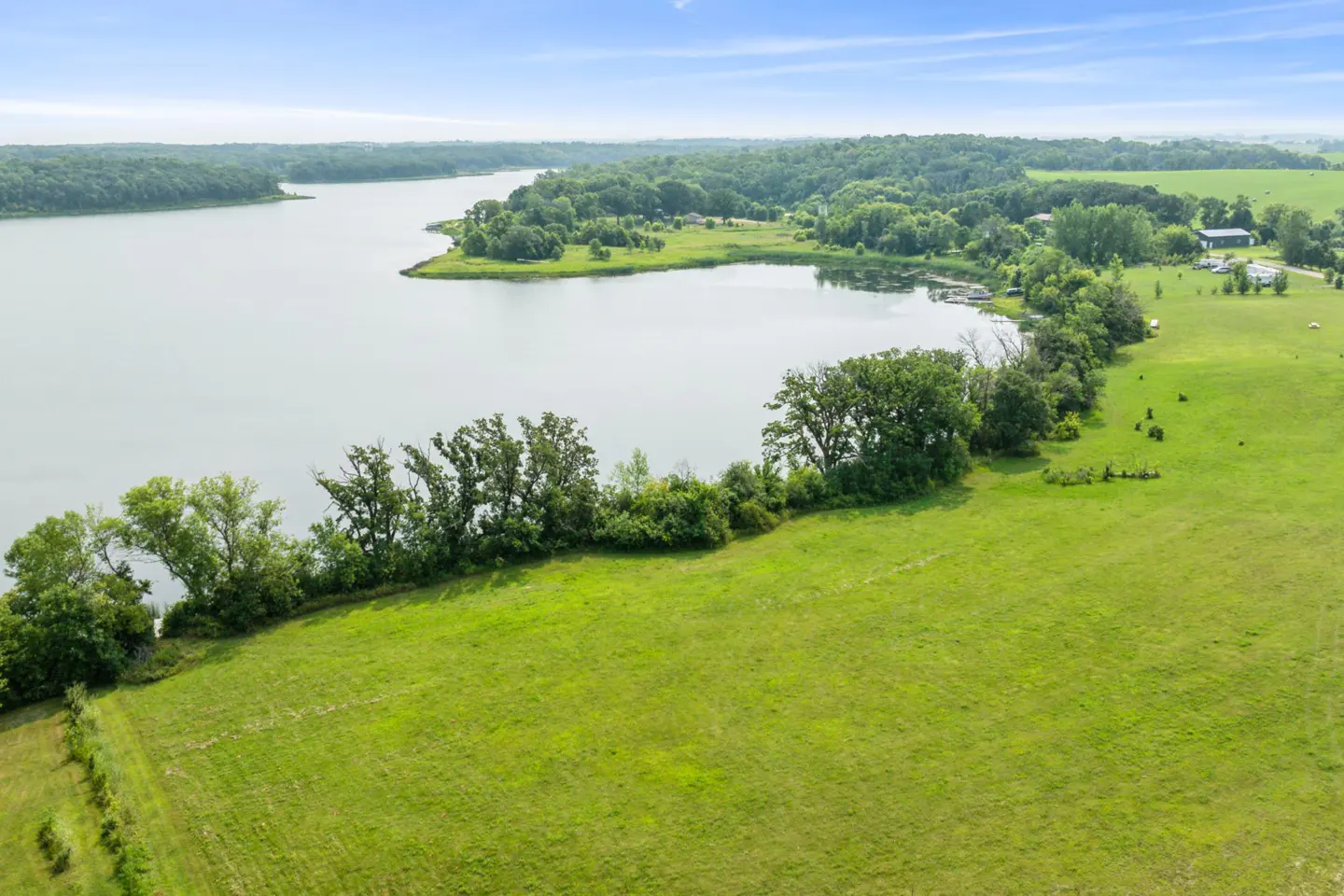 Aerial view of a lake surrounded by green fields and trees under a blue sky.