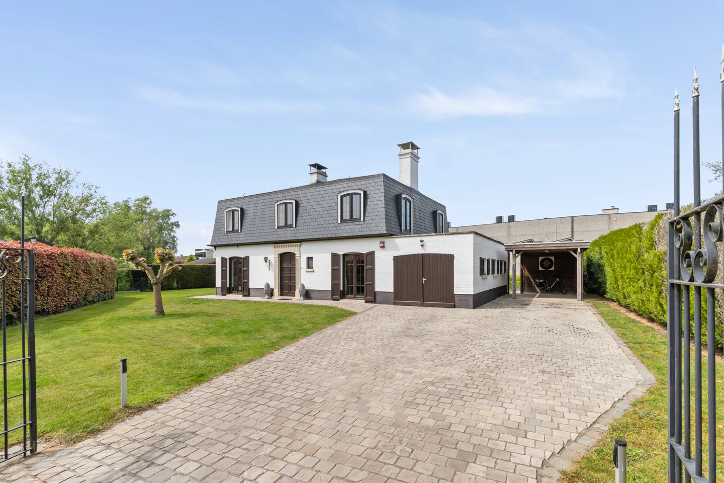 A white house with a gray roof and brown shutters sits behind a brick driveway and black metal gate.