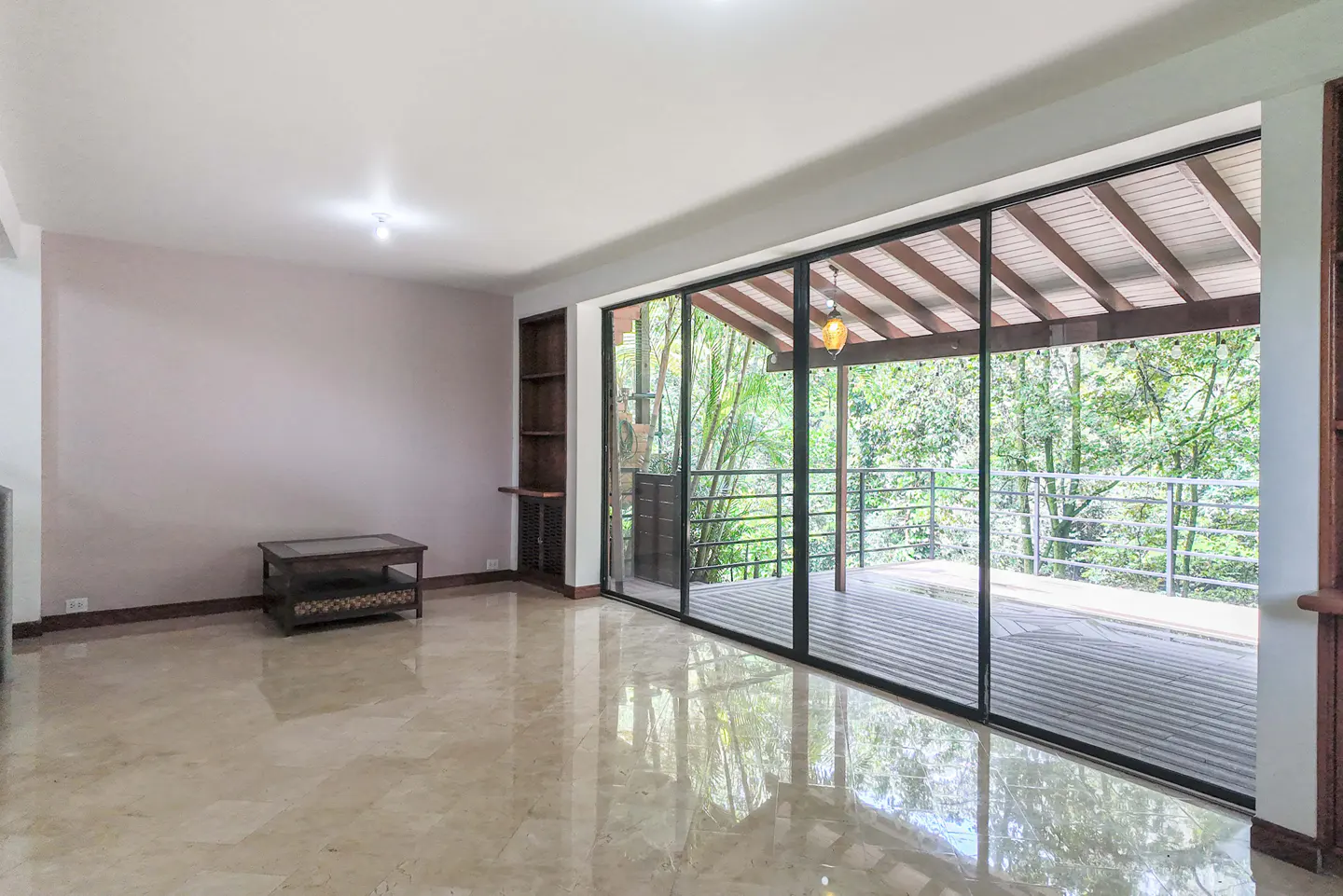 Bright living room with marble floors, a small table, and a large sliding glass door leading to a balcony overlooking lush greenery.