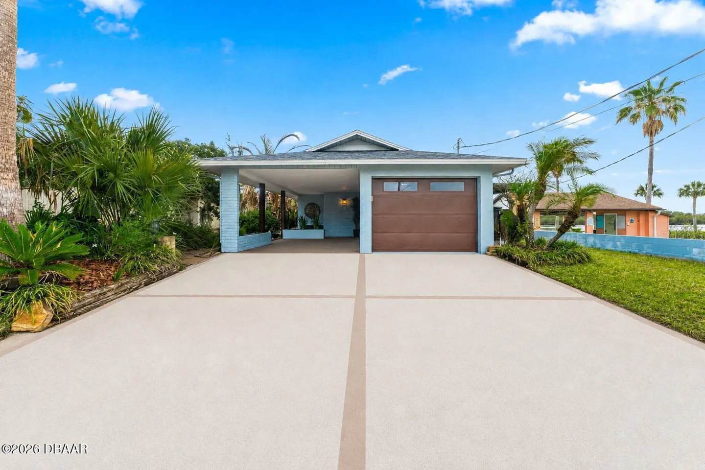 Front view of a light blue single-story house with a brown garage door and a long concrete driveway. Palm trees and blue sky in the background.