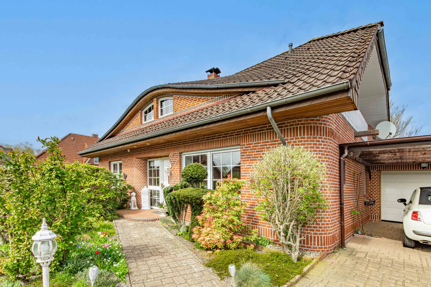 A red brick house with a brown tiled roof, a paved walkway, and a white car parked in the carport.