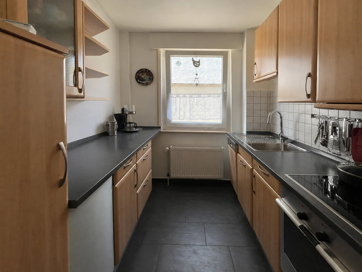 A narrow kitchen with light wood cabinets, dark countertops, and a black tile floor. A window is centered at the back.