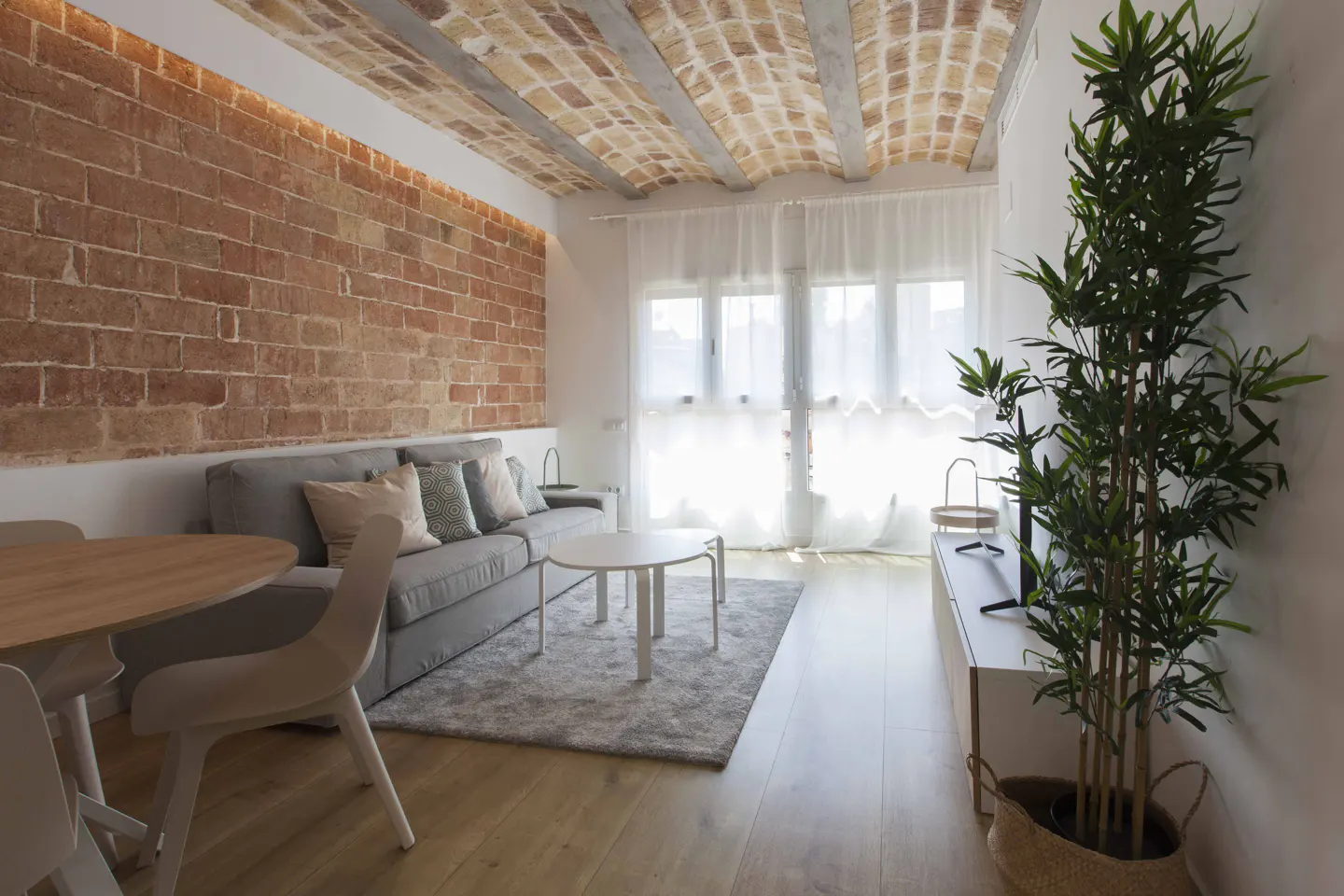 Modern living room with a brick wall, vaulted ceiling, grey sofa, round coffee table, and a tall potted plant.