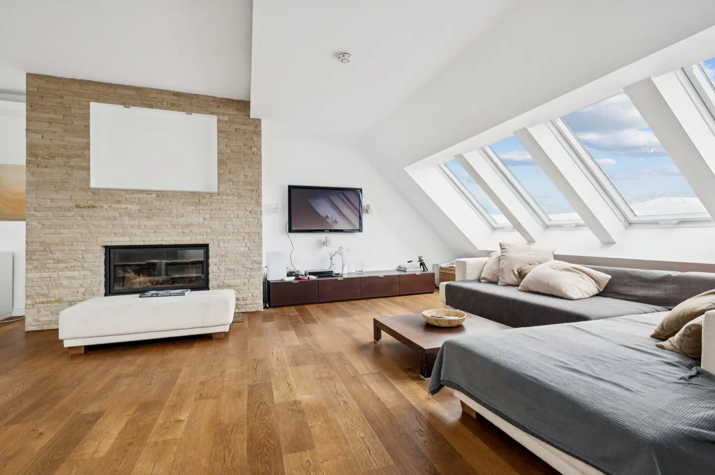 Bright living room with hardwood floors, a stone fireplace, and a gray sectional sofa under skylight windows.