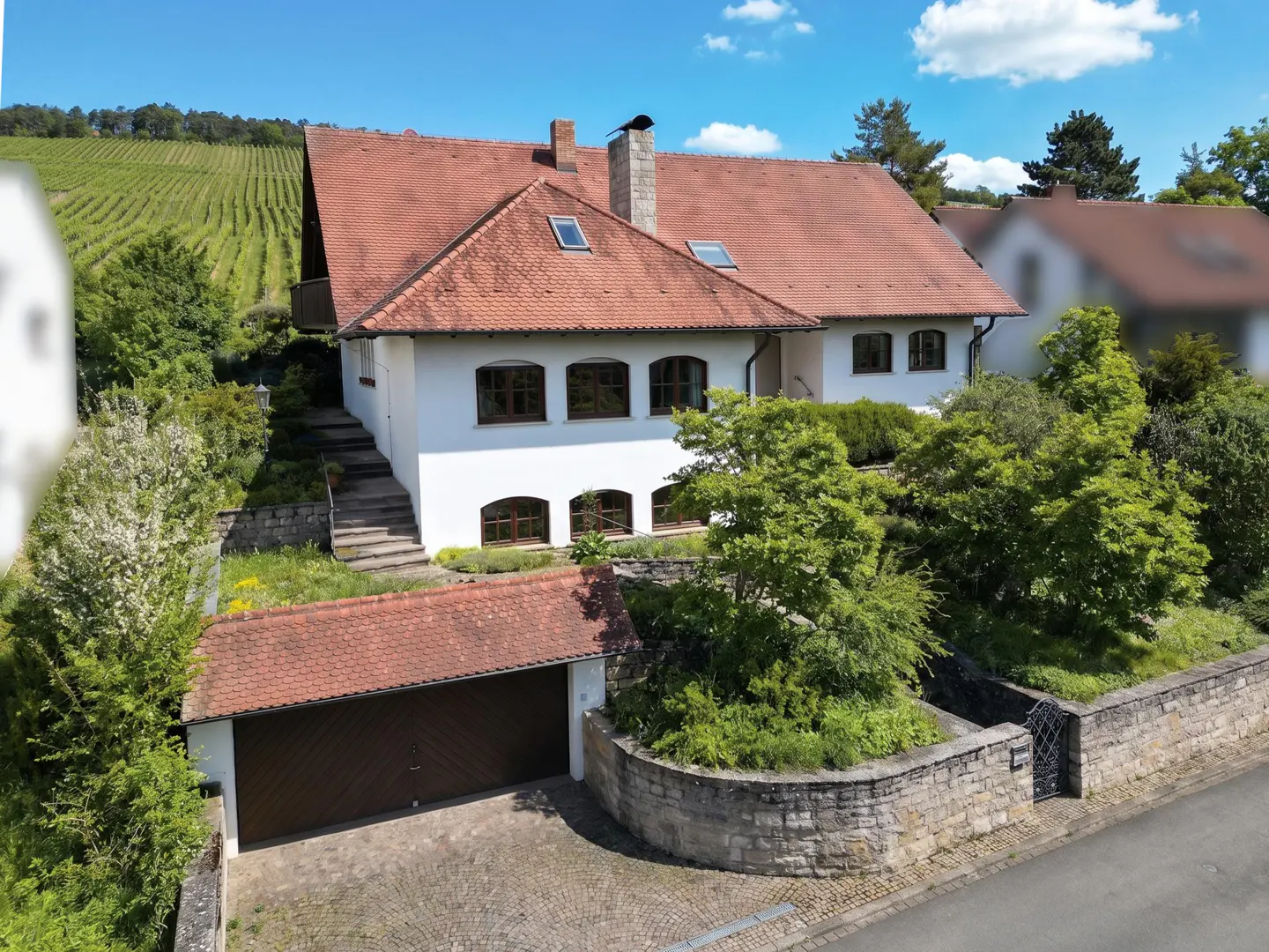 Two-story white house with a red tile roof, brown trim, and a brown garage door. A vineyard is visible in the background.