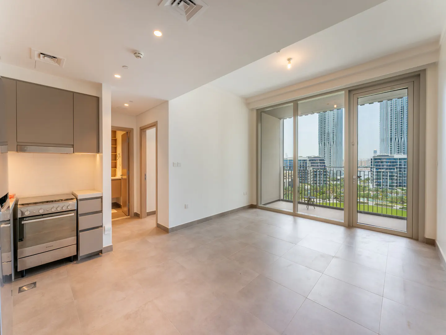 A bright, empty apartment with gray tile floors, a kitchen area with gray cabinets, and a balcony with a city view.