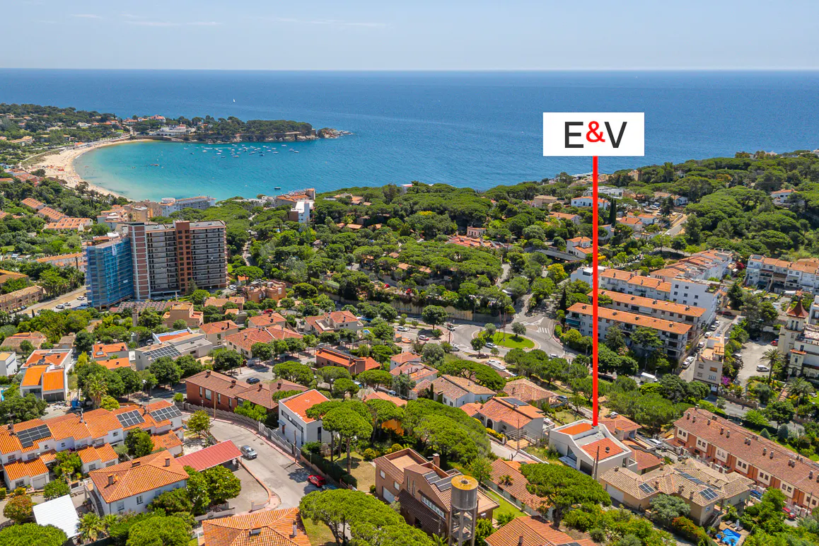 Aerial view of a coastal town with red-roofed houses, lush trees, and a sandy beach by the blue sea, marked by an E&V sign.